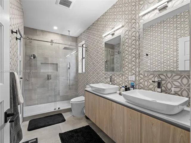 Bathroom featuring a dual-sink vanity with light wood-finish cabinetry, patterned wall tile, a frameless glass-enclosed shower with a rainfall showerhead, and wall-mounted faucets