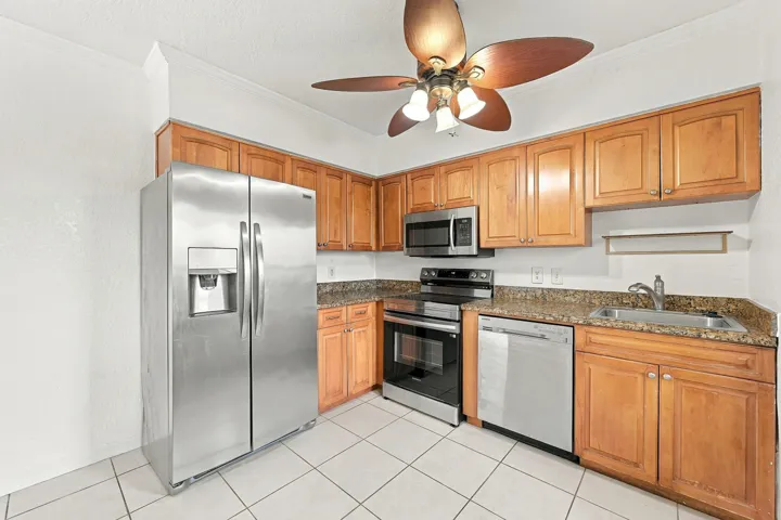 Kitchen featuring wood cabinetry, stone-finish countertops, stainless steel appliances, ceramic tile flooring, and a multi-blade ceiling fan with integrated lighting