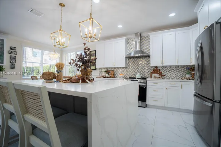 Kitchen featuring a waterfall-edge stone island, white shaker-style cabinetry, herringbone-patterned tile backsplash, stainless steel chimney-style range hood, and large-format tile flooring