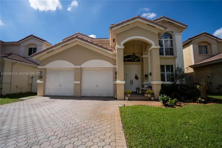 Two-story exterior featuring a stucco facade, tile roof, arched entryway with decorative columns, and a paved stone driveway leading to an attached two-car garage