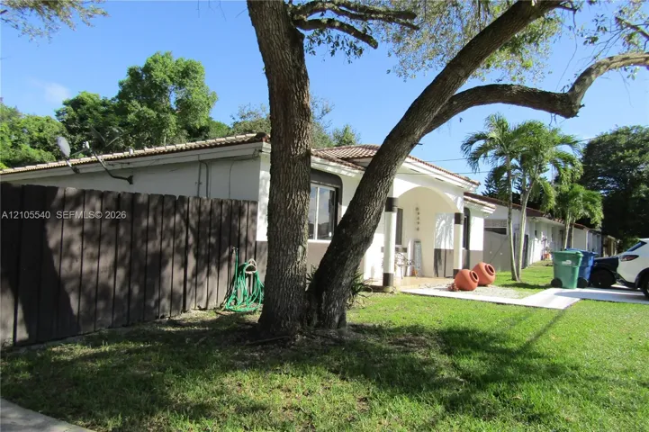 Manicured front lawn with mature trees, a white stucco exterior, dark wood fencing, and a tiled roof