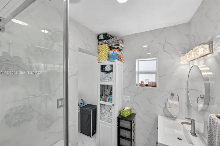 Full-height, large-format stone-patterned wall tile, glass-enclosed shower, round wall-mounted mirror, integrated rectangular sink with metallic faucet, and multi-bulb vanity lighting