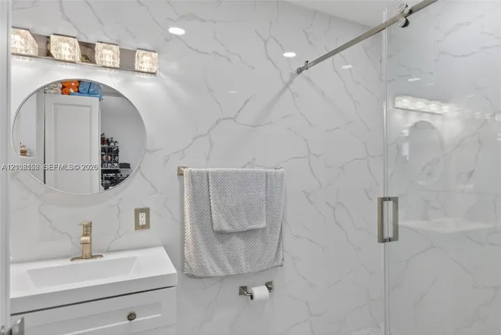 Bathroom featuring large-format wall tiling with veining patterns, a vanity with an integrated rectangular sink, a circular mirror, a glass-enclosed shower with metal hardware, and a four-light fixture