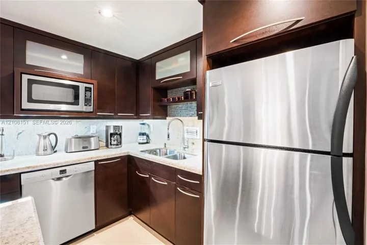 Kitchen featuring dark wood cabinetry, stainless steel appliances, light-toned stone countertops, and a glass tile backsplash