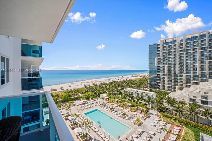 Private balcony with glass panel railing overlooking an outdoor swimming pool, landscaped grounds, and coastal shoreline