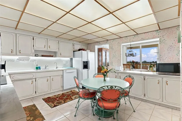 Kitchen featuring light-finish cabinetry, under-cabinet task lighting, white tile flooring, a geometric grid ceiling, and a built-in countertop extension