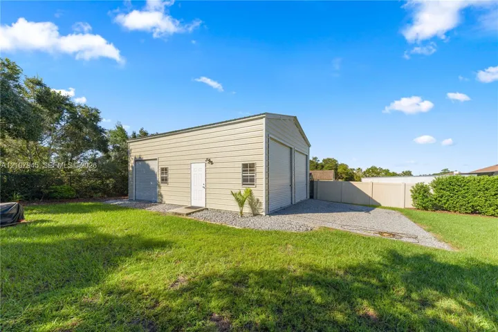 Detached metal outbuilding featuring a high-clearance garage door, a side-entry personnel door, dual windows, and an expansive gravel driveway