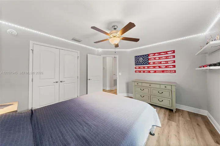 Room featuring wood-finish flooring, recessed perimeter LED accent lighting, a multi-blade ceiling fan with integrated light fixture, double-door closet, and white floating wall shelves