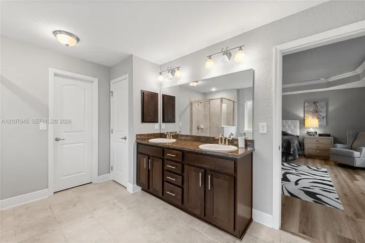 Dual-sink vanity with dark wood cabinetry, stone-finish countertop, full-width vanity mirror, and tile flooring