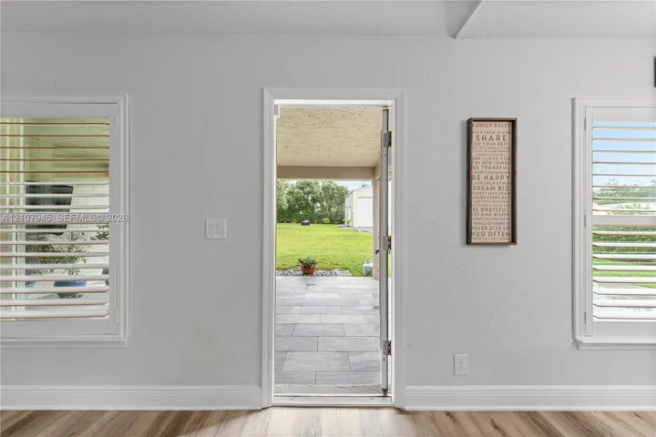 Interior featuring wood-finish flooring, dual windows with plantation-style shutters, and an exterior door opening to a paved patio area