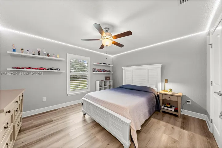Wood-finish flooring, ceiling fan with integrated lighting, window with shutter treatments, and installed wall-mounted shelving