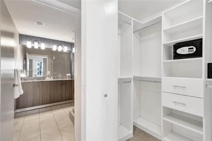 Walk-in closet featuring white built-in shelving, drawers, and hanging rods, accompanied by a bathroom with a dual-sink vanity, stone-finish countertops, tile flooring, and wall-mounted light fixtures