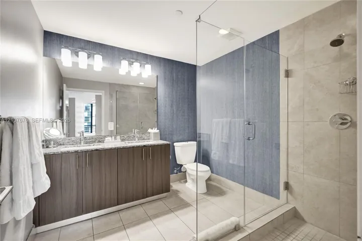 Bathroom featuring a dual-sink vanity with stone countertop, frameless glass-enclosed shower, large-format tile flooring, and integrated wall lighting