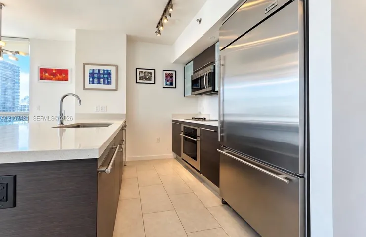 Kitchen featuring dark wood-finish cabinetry, stone-composite countertops, stainless steel appliances, a center island with an undermount sink, and tile flooring
