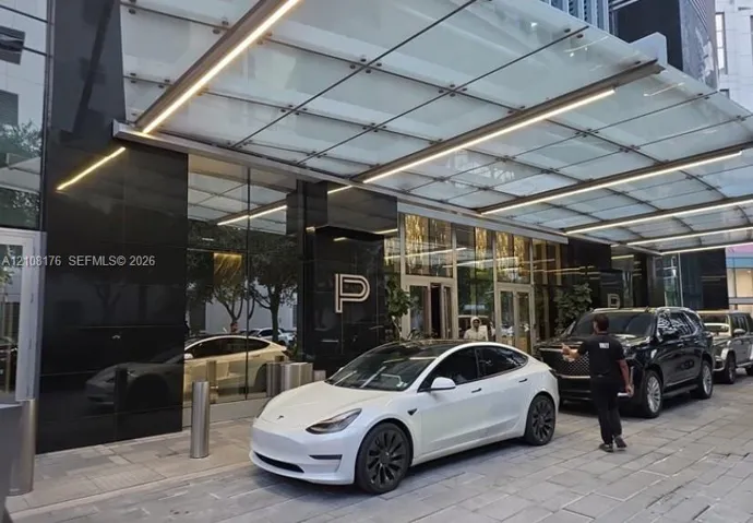 Covered porte-cochère featuring a glass canopy with integrated linear lighting, polished black stone facade, and stone-paved motor court