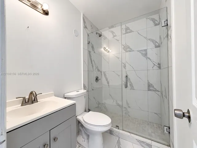 Bathroom featuring a grey shaker-style vanity with an integrated sink, walk-in shower with large-format stone-patterned tile, frameless glass enclosure, and modern brushed-metal fixtures