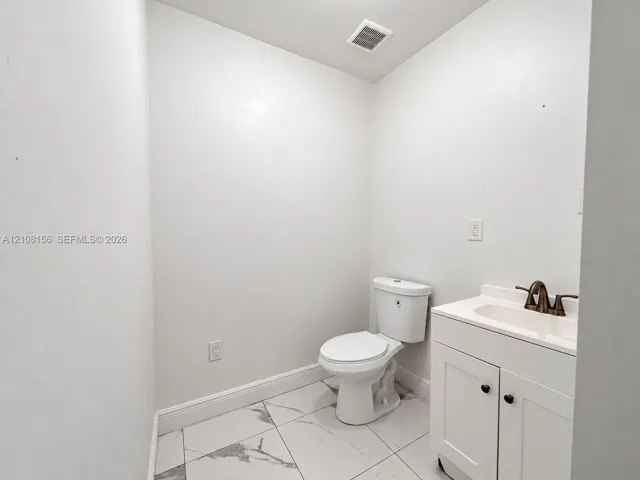 White vanity with a neutral countertop, dark metal faucet, ceramic toilet, and large-format tile flooring with a veined pattern