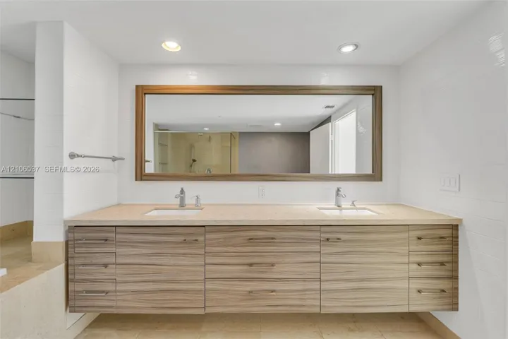 Floating vanity featuring wood-grain cabinetry, dual undermount sinks, stone-surface countertop, and a large framed mirror with integrated recessed lighting