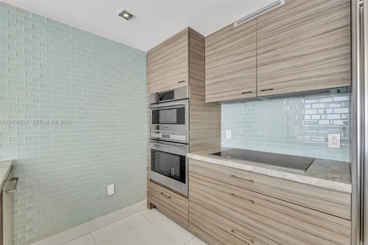 Modern kitchen featuring wood-finish cabinetry, light blue subway tile backsplash, built-in stainless steel double oven, and integrated cooktop