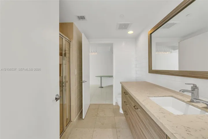 Bathroom vanity with neutral stone-style countertop, wood-finish cabinetry, rectangular undermount sink, and full-width framed mirror, complemented by a glass-enclosed shower featuring textured tile surround