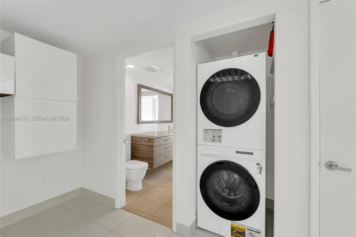 Laundry area featuring a stacked washer and dryer unit, built-in cabinetry, ceramic tile flooring, and a doorway to a bathroom equipped with a wood-finish vanity and rectangular mirror