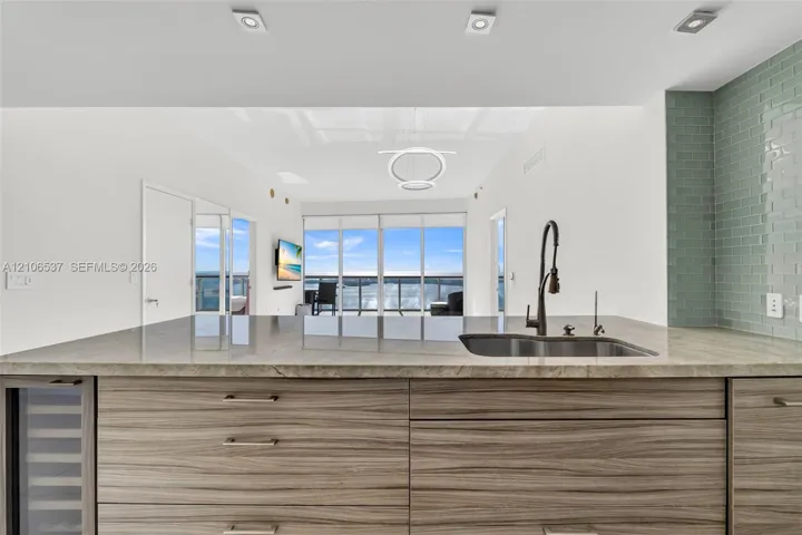 Kitchen island featuring wood-finish cabinetry, stone countertop, undermount sink with a matte black pull-down faucet, and a built-in wine cooler, complemented by a light green subway tile backsplash