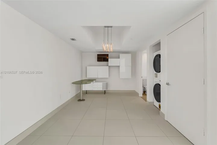 Modern utility space featuring large-format tile flooring, built-in glossy white cabinetry, an integrated stone-surface workstation, a tiered pendant light fixture, and a stacked washer and dryer unit