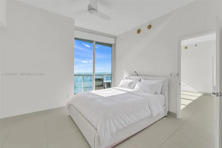 Large-format tile flooring, ceiling fan with integrated lighting, and floor-to-ceiling glass sliding doors leading to a private balcony with water scenery