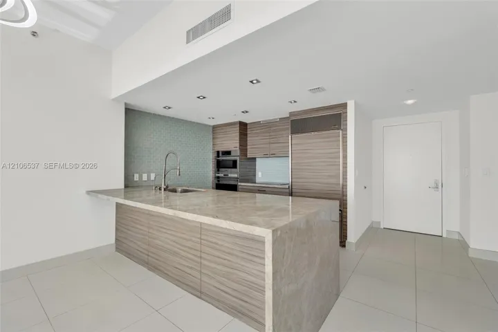 Kitchen featuring a waterfall-edge stone countertop, wood-finish cabinetry, built-in stainless steel refrigerator, recessed lighting, and patterned tile backsplash