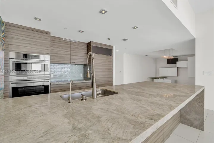 Kitchen featuring a waterfall-edge stone countertop, wood-finish cabinetry, stainless steel built-in double wall oven, recessed lighting, and a dual-basin stainless steel sink with a high-arch faucet