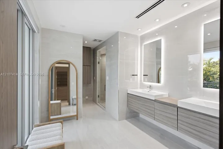 Dual floating vanity with horizontal wood-finish cabinetry, integrated rectangular sinks, and backlit mirrors, accented by large-format neutral wall tiles and recessed lighting