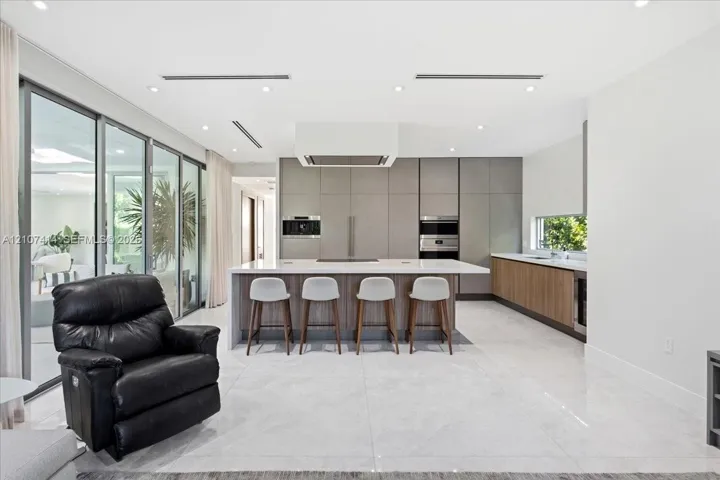 Chef’s kitchen featuring full-height matte cabinetry, a waterfall-edge stone island, built-in stainless steel appliances, polished stone flooring, and expansive floor-to-ceiling glass sliding doors
