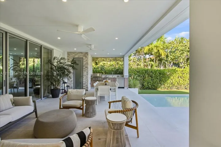 Covered outdoor patio featuring light-toned tile flooring, recessed ceiling lighting, ceiling fans, and a built-in outdoor kitchen with an integrated grill and stone-style backsplash, overlooking a private swimming pool