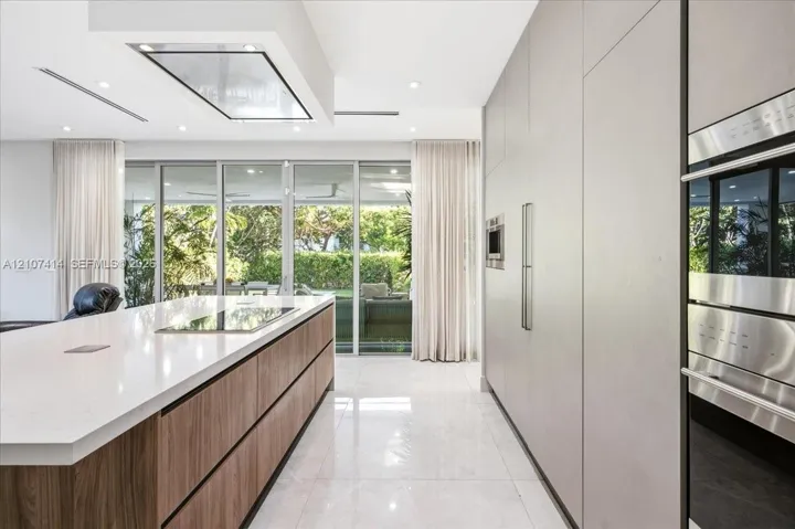 Contemporary kitchen featuring a large island with wood-finish cabinetry, a waterfall-edge stone countertop, integrated flush-panel cabinetry, stainless steel built-in ovens, and floor-to-ceiling glass sliding doors