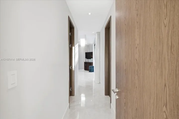 Hallway featuring large-format light-toned stone flooring, recessed ceiling lighting, and solid wood-finish doors with brushed-metal hardware