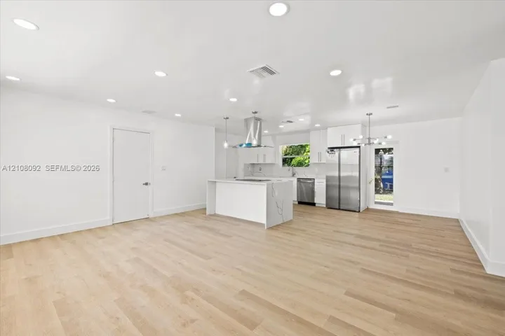Open-concept interior featuring light wood-finish flooring, recessed lighting, a central island with stone-finish waterfall countertop, stainless steel appliances, and white shaker-style cabinetry