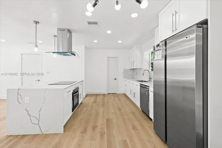 Kitchen featuring white cabinetry, wood-finish flooring, a waterfall-edge stone island with a built-in oven, stainless steel appliances, and a modern chandelier