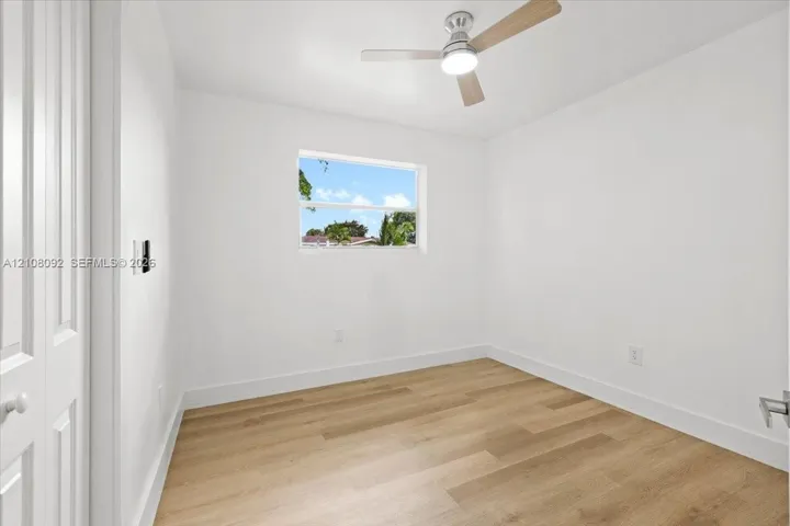 Neutral-toned room featuring wood-finish flooring, a ceiling fan with integrated lighting, a single-pane window, and white interior doors