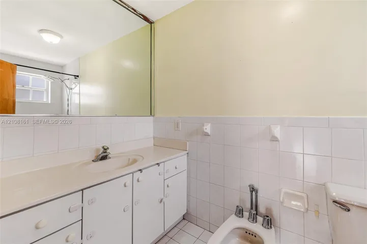 Bathroom featuring a white vanity with an integrated sink, a full-width wall mirror, white wall tiling, and a bidet