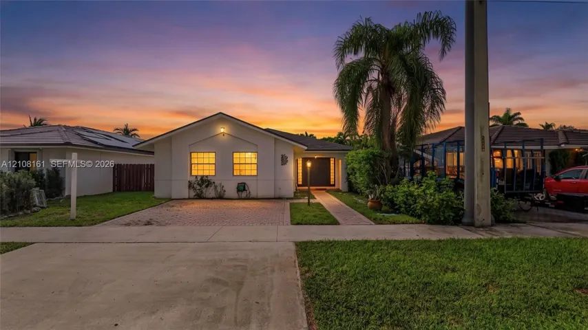 Single-story exterior featuring a light-toned stucco façade, paver driveway, covered entryway, and mature palm tree