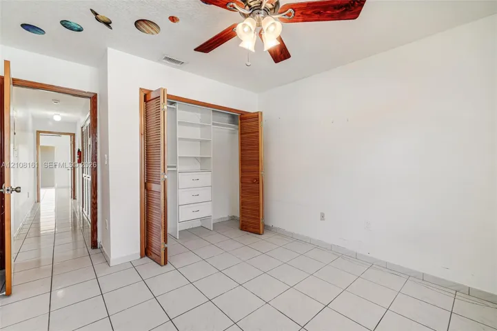 Ceramic tile flooring, built-in closet storage system with integrated shelving and drawers, wood-finish louvered bi-fold closet doors, and ceiling fan with integrated lighting