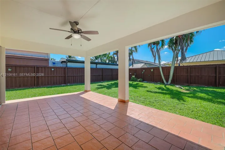 Covered patio featuring terra-cotta tile flooring, a ceiling fan, a fenced perimeter, and a manicured lawn