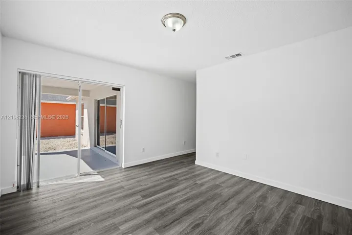 Interior space featuring wood-finish flooring, white walls, flush-mount ceiling lighting, and sliding glass doors with vertical blinds leading to a paved patio area