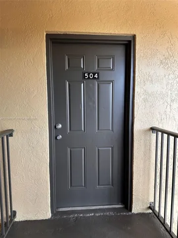 Six-panel entry door finished in a dark neutral tone, featuring a dual-lock handle set and numerical address plaque, framed by textured exterior stucco walls