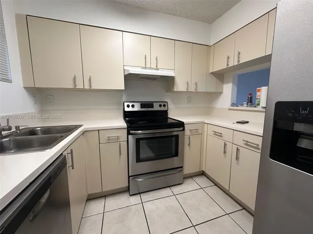 Kitchen featuring cream-toned cabinetry with metal hardware, solid-surface countertops, stainless steel appliances, a dual-basin sink, and tile flooring