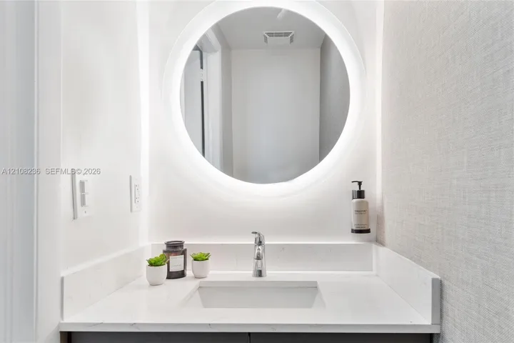 Circular backlit wall mirror, stone-composite countertop with integrated sink, chrome single-lever faucet, and textured wall finish
