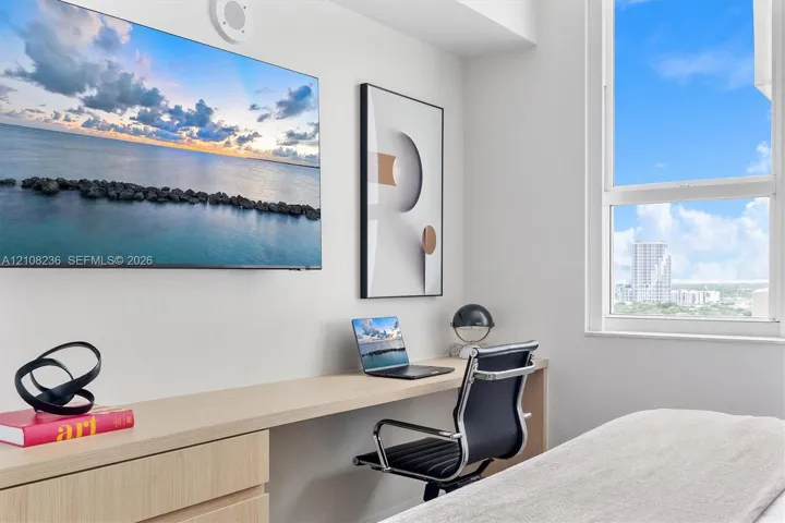 Custom built-in desk with light wood-finish cabinetry, ceiling-mounted speaker, and large-format window featuring expansive sky and urban scenery