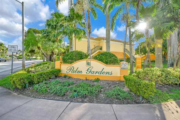 Stucco exterior community signage featuring masonry pillar accents, decorative landscaping, and mature palm trees