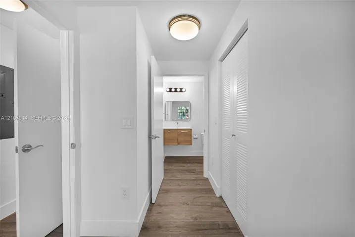 Hallway featuring wood-finish flooring, white louvered closet doors, and a flush-mount ceiling light fixture, opening into a space with a wall-mounted vanity, rectangular mirror, and four-light vanity bar
