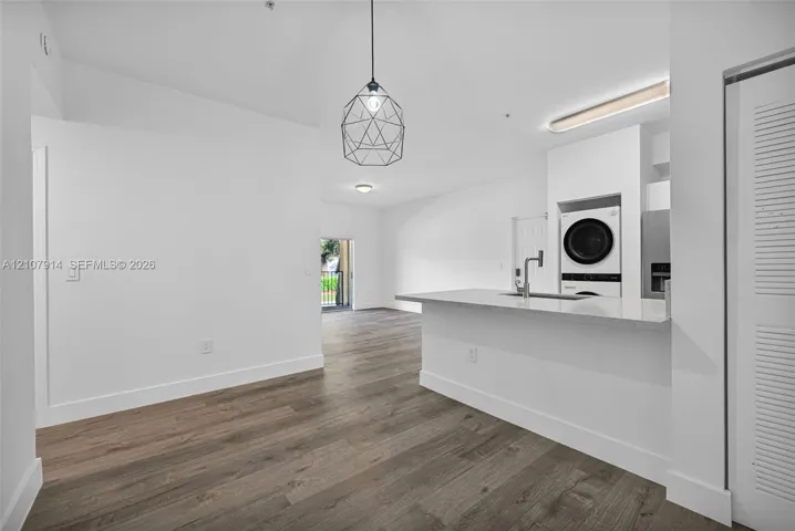 Kitchen featuring a quartz countertop island with an integrated sink, geometric pendant lighting, wood-finish flooring, and built-in stacked laundry appliances